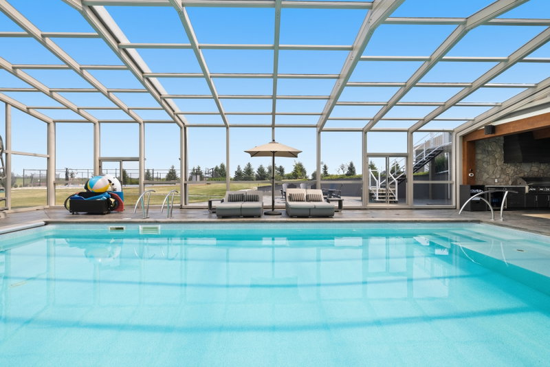 Glass-enclosed indoor pool with lounge seating and an outdoor kitchen beyond